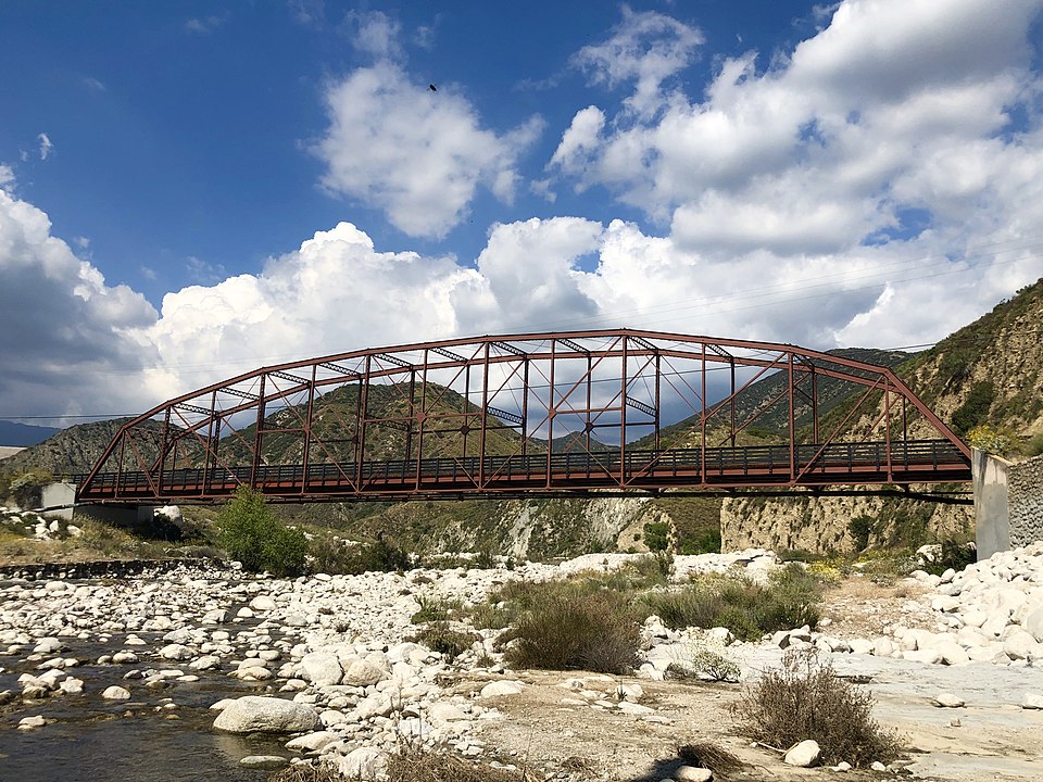 Santa Ana River Bridge, a Red iron truss bridge spanning a river with rocky banks under a partly cloudy sky, symbolizing the efficiency and sustainability akin to adopting Inland Empire Solar solutions for cheaper electricity.