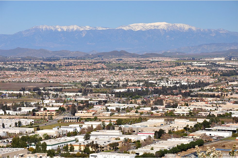 A panoramic view of a sprawling suburban area with low-rise buildings, featuring solar energy solutions, bordered by distant mountains capped with snow under a clear blue sky.