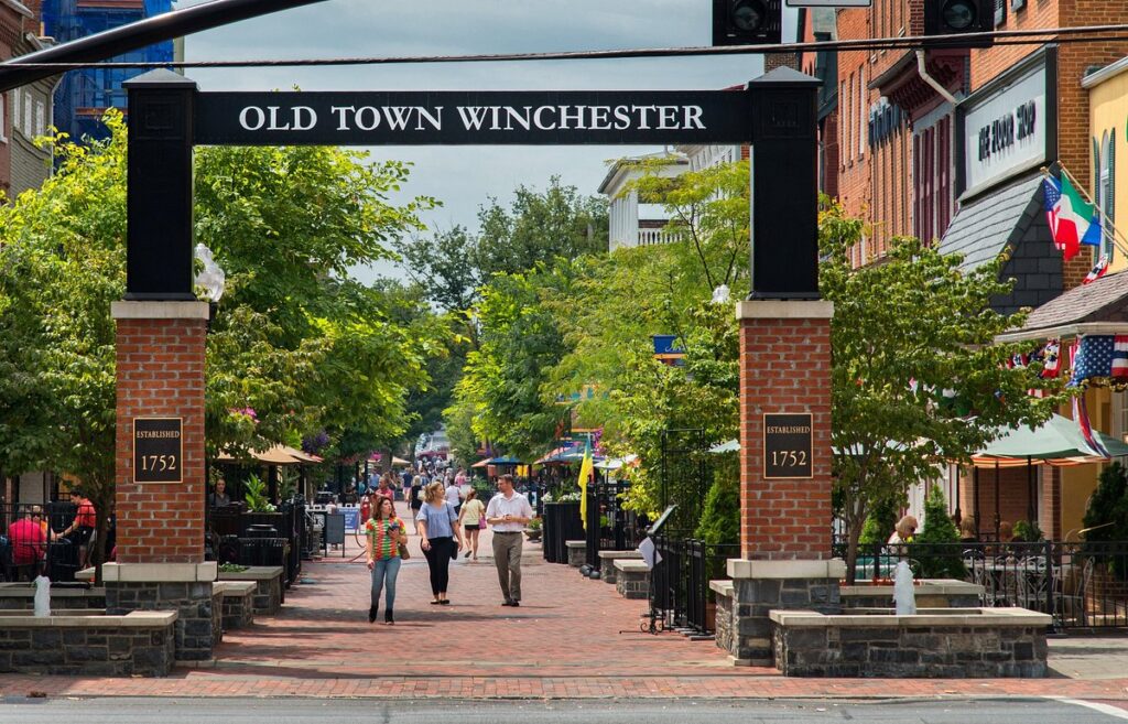 A vibrant street scene in old town Winchester, CA, featuring people walking, brick-lined sidewalks, multicolored flags, and diverse storefronts under a prominent archway reading "Old Town Winchester.